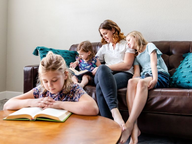 Mother on brown leather couch reading with a toddler and older child, while a middle child reads at a coffee table in front.