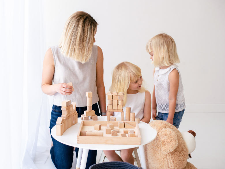 A blonde mother playing blocks with her two blonde daughters.