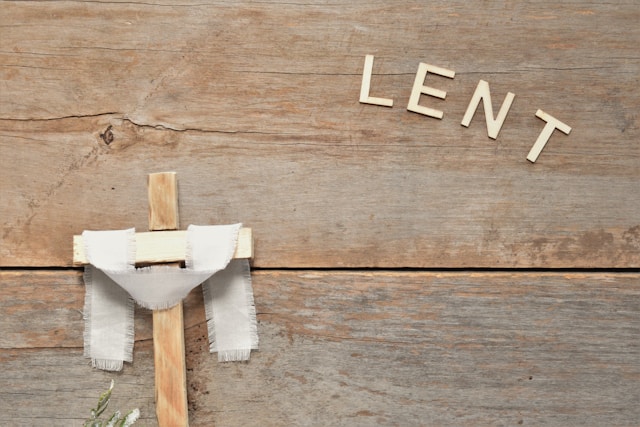 Small wooden cross with a white cloth draped over it hung on wooden wall bearing the word "LENT" in the top righthand corner.
