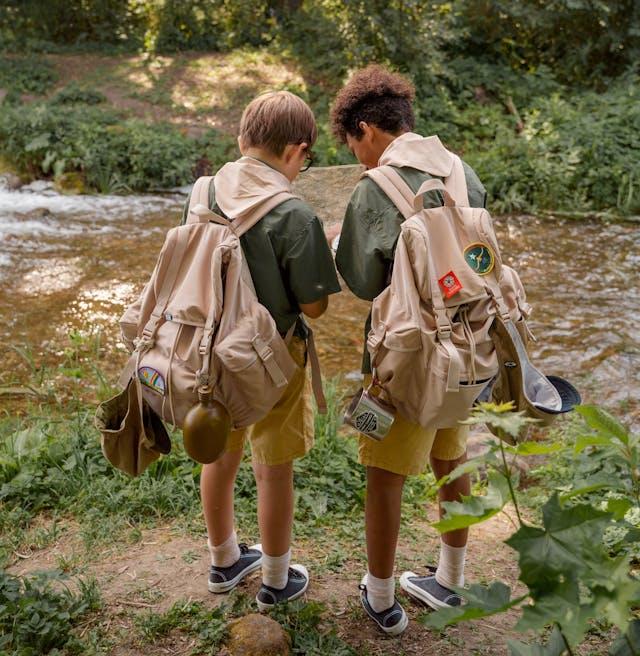 Back view of two Boy Scouts standing by a stream full backpacks with their heads together.