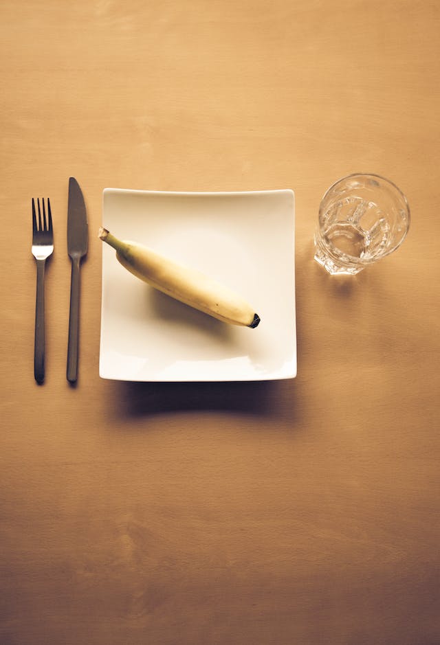Square white plate on a table with one banana on it, fork and knife to the left side, and a glass or water next to the top right corner
