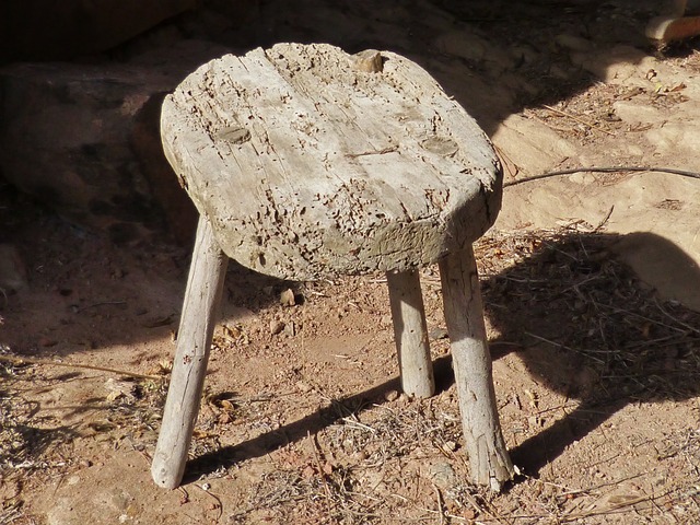 Old three-legged stool sits on the ground casting a shadow.