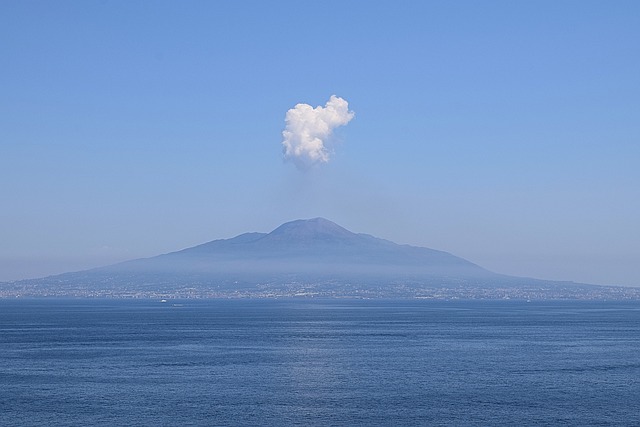 Volcano with plume of smoke rising from the top as seen across a body of water. 