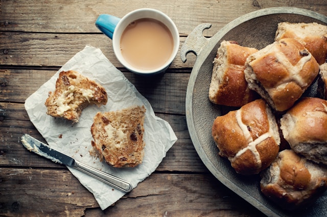 Coffee cup with hot cross buns to right and knife lying next to plate with split bun which has been buttered