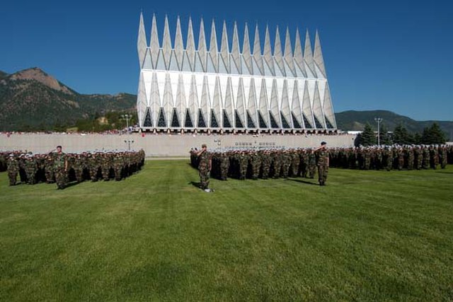 Cadets in camouflage uniform stand in formation in the grass with the Cadet Chapel in the background