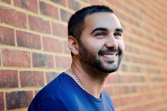 Bearded man in blue shirt seen from the shoulders up stands in front of brick wall smiling 
