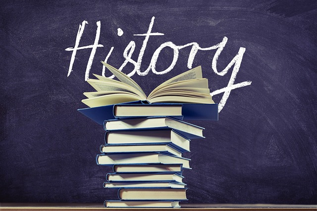 Books stacked in a pile with the top book open and a blackboard with "History" written on it behind them