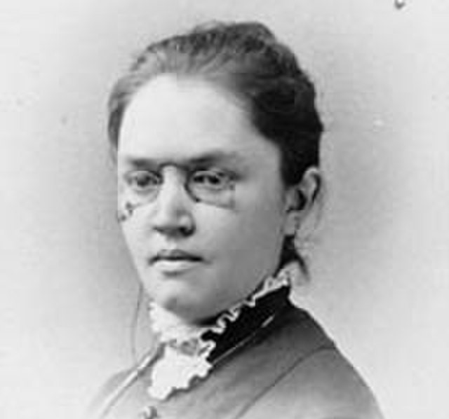 Black and white photographic portrait of a woman from the shoulders up wearing pince-nez glasses and with her hair in a bun