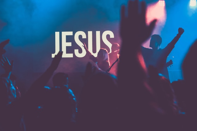 View of concert in progress through crowd of raised hands with the word "JESUS" displayed behind the band