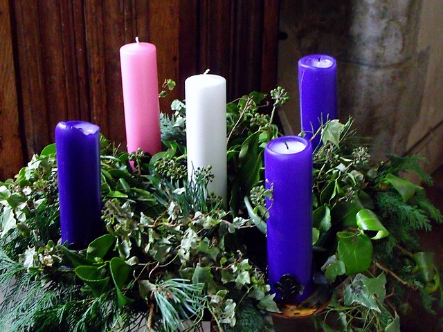 Advent wreath with five candles set in greenery. A white candle is in the middle and three purple and one pink candle surround it.