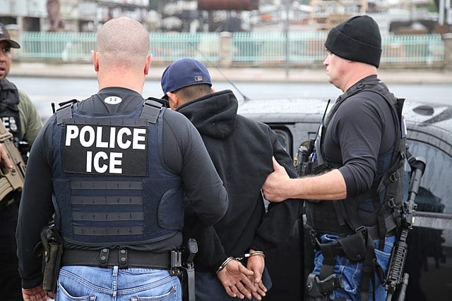 View from behind of three men from waist up with the one in the middle's hands handcuffed behind his back and the two mean flanking him with the one on the left wearing a shirt saying "POLICE ICE." 