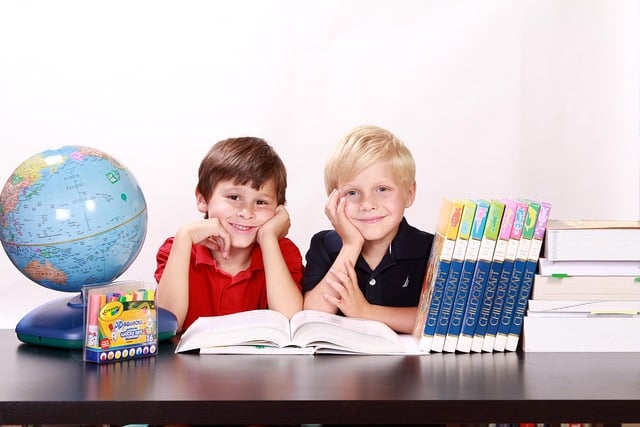 Two smiling young boys sitting side-by-side in a classroom setting with an open book in front of them