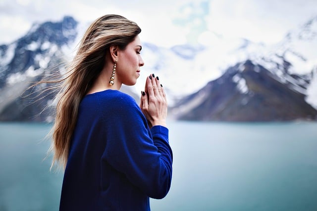 Side view of woman with long hair in purple sweater and hands clasped in prayer before her face with a lake and mountains in the background