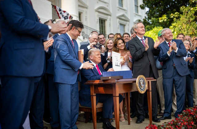 President Donald seated surrounded by standing individuals as he holds up a bill he just signed