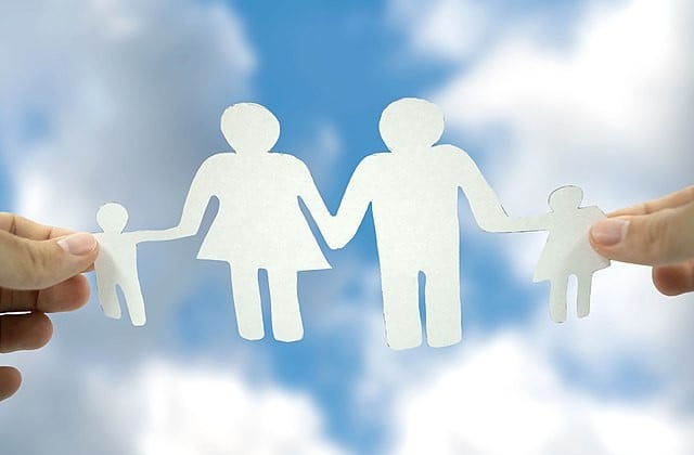 Paper cutouts of mother, father, and two children held up by a hand on each end against a blue sky with clouds
