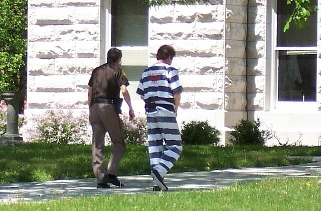 View from behind of officer escorting prisoner in black and white striped uniform towards a building 