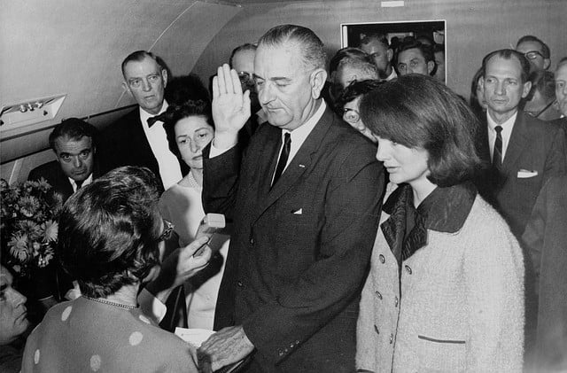 Black and white photo of Lyndon Johnson in a suit with his right hand raised taking the oath of office as President of the United States with Jackie Kennedy standing beside him