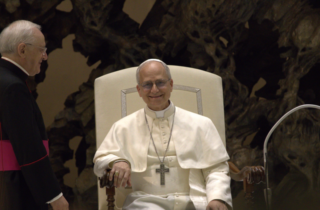 Pcitrue of a smiling Pope Leo sitting in a white, high=backed chair in a white clerican garment and a crucifix on his chest from a chain around his neck