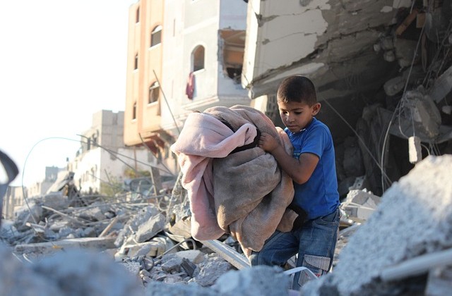 Young boy in blue short-sleeve shirt and jeans sitting on rubble holding bedding