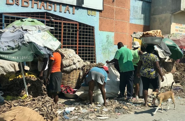 People picking through trash in a Haitian street in front of a dilapidated building