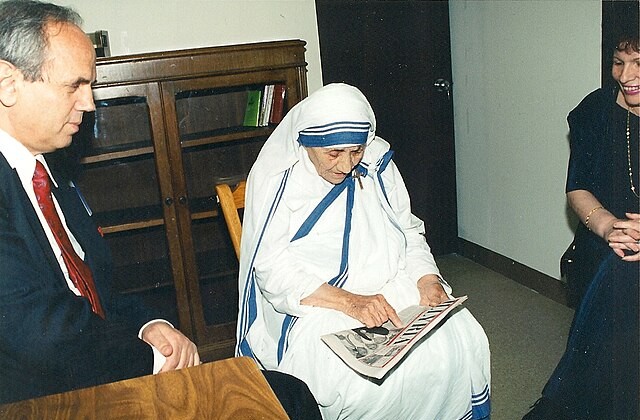 View of Mother Theresa in a room seated in a chair with a newspaer in her lap. A man and a woman sit just off to opposite sides of her.