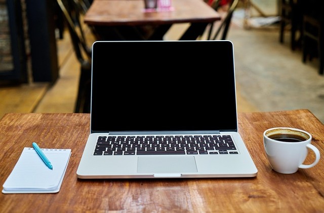 Open laptop sitting on a wooden table with a coffee mug to the right and a pad and pen to the left 
