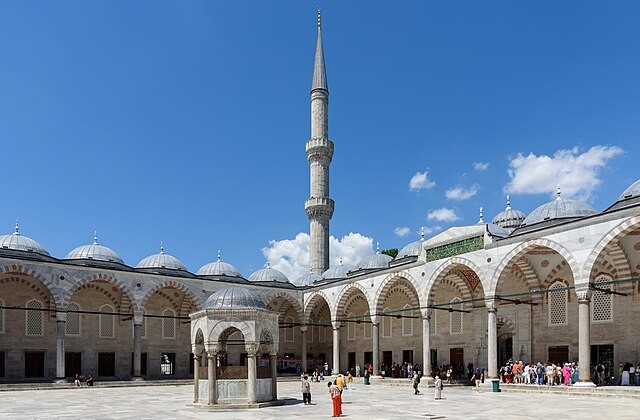 View from the corner of the courtyard of Istanbul's Blue Mosque. A minaret rises into a blue sky with white clouds in the center of the picture. Visitor can be seen in various spots at this site.
