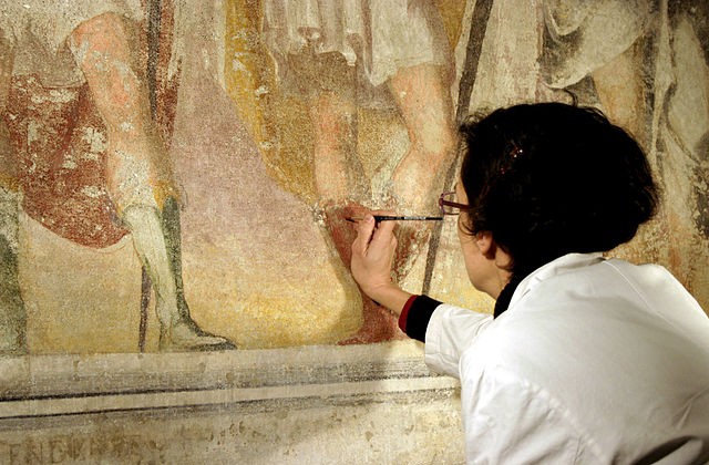Person with back to camera wearing white coat and glasses with paintbrush in hand working on artwork