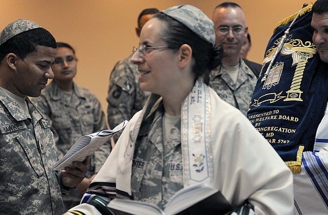 View from waist up of uniformed female Jewish chaplain wearing a prayer shawl and glasses and holding an open book.