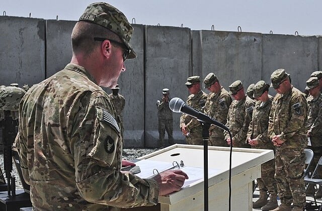 Man standing at podium speaking into microphone wearing camouflage and facing similarly attired men with heads bowed.