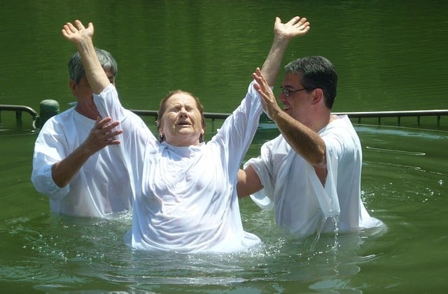 Woman in river wearing white and holding her arms up as she is baptized by two people also in white on either side of her
