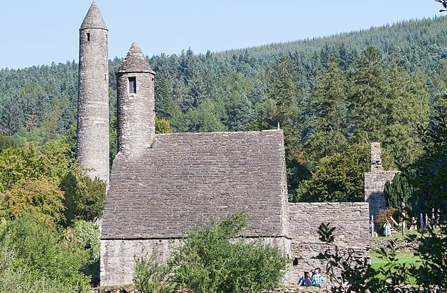 Side view of stone church with round tower in the background in a wooded setting.
