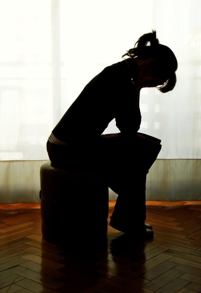 Silhouette of a woman sitting alone in a room with her head bent down and hands covering her face.