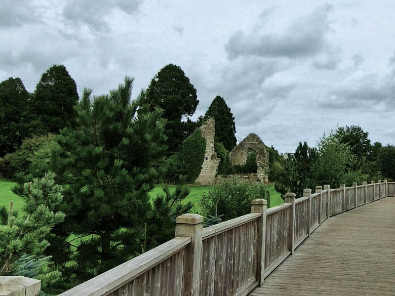 Image of a wooden bridge surrounded by vibrant greenery and what appears to be a small ruin. 
