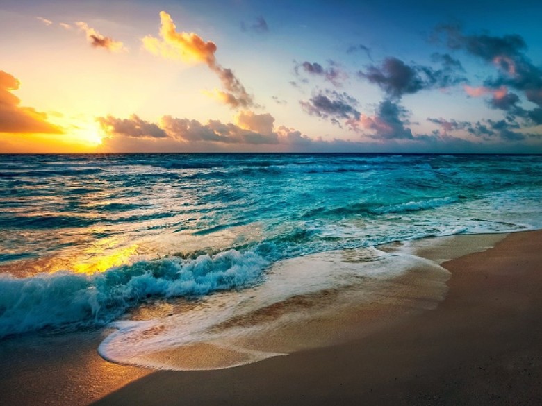 Sunset over turquoise colored ocean with gentle waves rolling onto a sandy shore, The sun reflects off the water as the light foam from the tide roll into the shoreline.