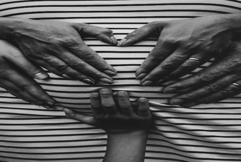 The image captures the meaning of motherhood as the hands of a wife and mother around her womb in the shape of a heart. The hands of a husband and father are on the outer layer of the wife's hands while a child's hands are placed on the bottom center of the womb. 
