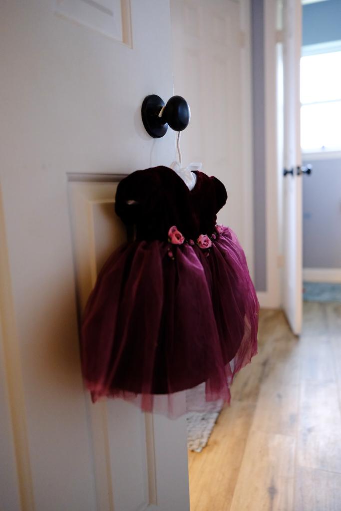 Maroon baby dress hanging on a hanger on a doorknob in a room with natural light