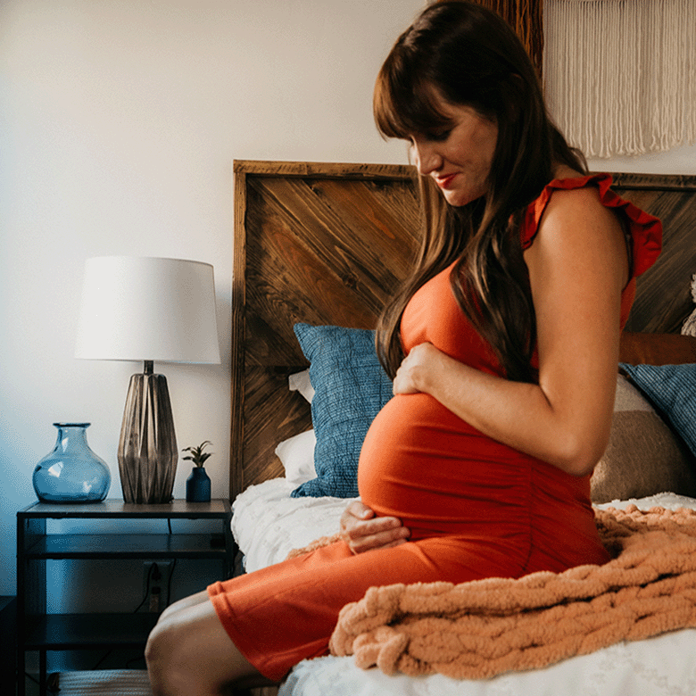 pregnant woman in your 40s in an orange dress sitting on a bed and holding her belly