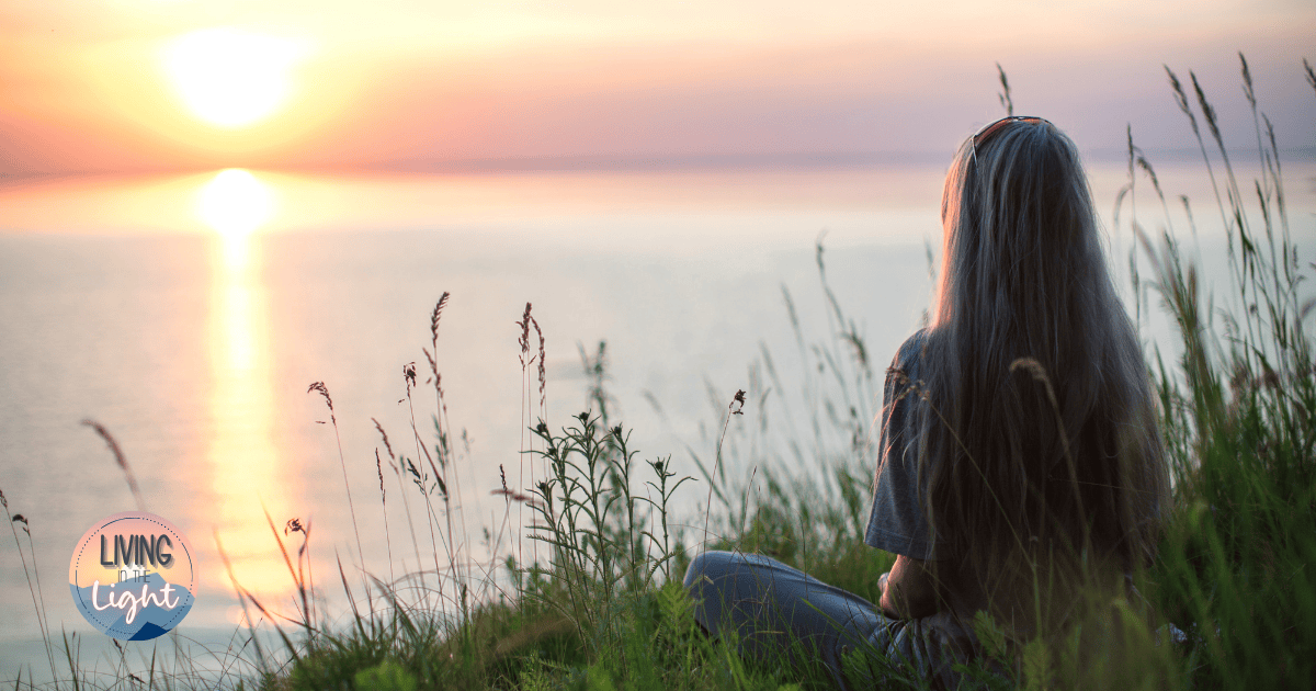 Woman sitting in tall grass at sunset, reflecting quietly overlooking the water, symbolizing mental rest, peace, and emotional wellness.