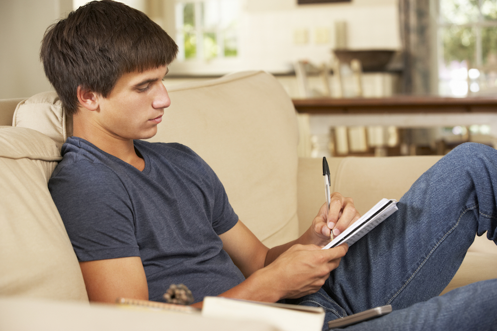 Teenage Boy Sitting On Sofa At Home Doing Homework