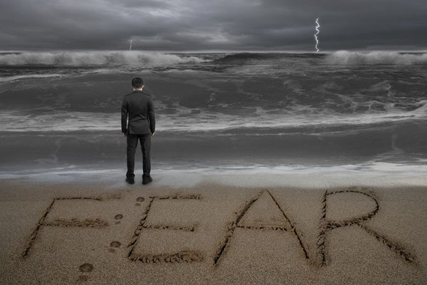 39296466 - rear view of black suit businessman standing facing fear word written on sand beach dark stormy ocean background