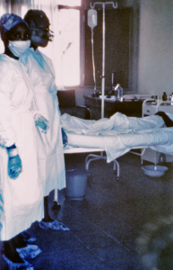 Two nurses standing in front of Ebola case #3, who was treated, and later died at Ngaliema Hospital, in Kinshasa, Zaire. (1976, public domain)