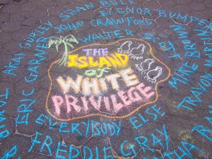 Image: "The Island of White Privilege, written in chalk with a palm tree and footprints, surrounded by the names of Black people killed by police.