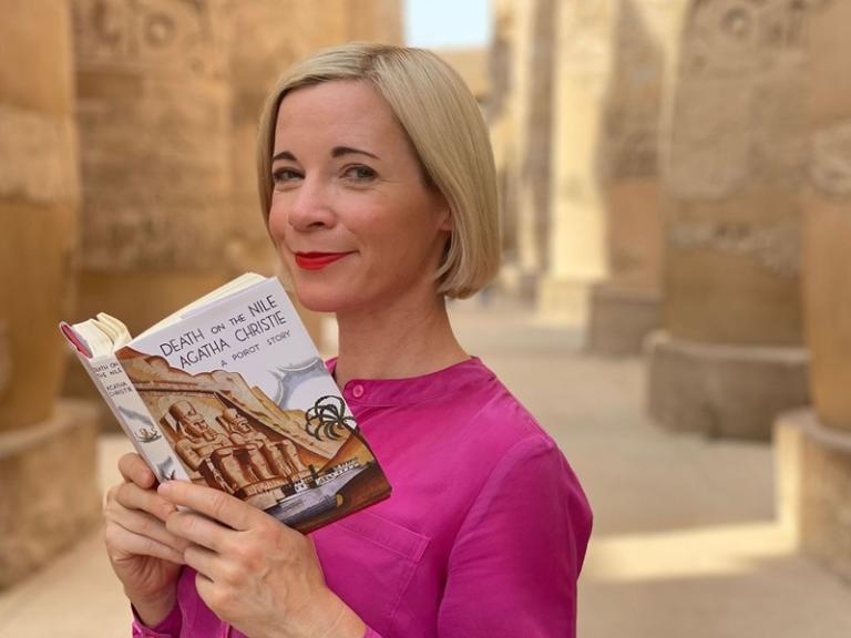 A blonde woman in a pink dress reads an Agatha Christie novel in an Egyptian temple.