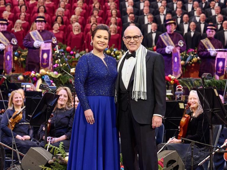 A woman and man in festive evening dress stand in front of a choir and orchestra.