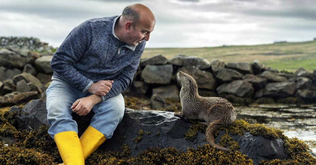 A man and an otter sit together on a rocky shoreline.