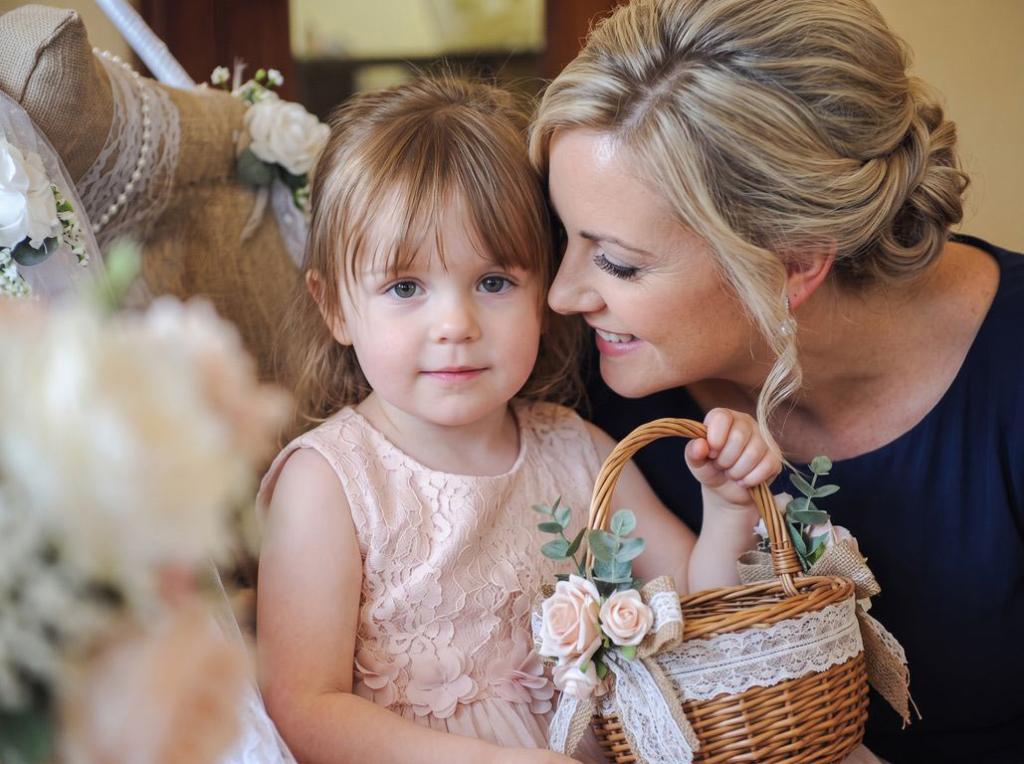 A tender, candid photograph of a blonde mother and her young daughter at a formal event. The little girl in a blush-pink lace dress holds a flower basket decorated with ribbon and greenery, while her mother smiles warmly as she leans in close to talk with her.