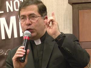 Frank Pavone in a Roman collar, yakking into a microphone with one finger upraised.