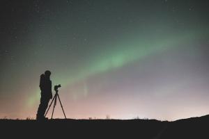 A person with a camera in silhouette against a night sky with a green and blue aurora 