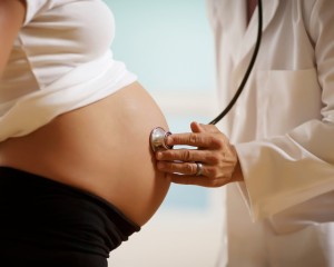 Pregnant woman with male doctor to woman abdomen in exam room to listen to heartbeat.